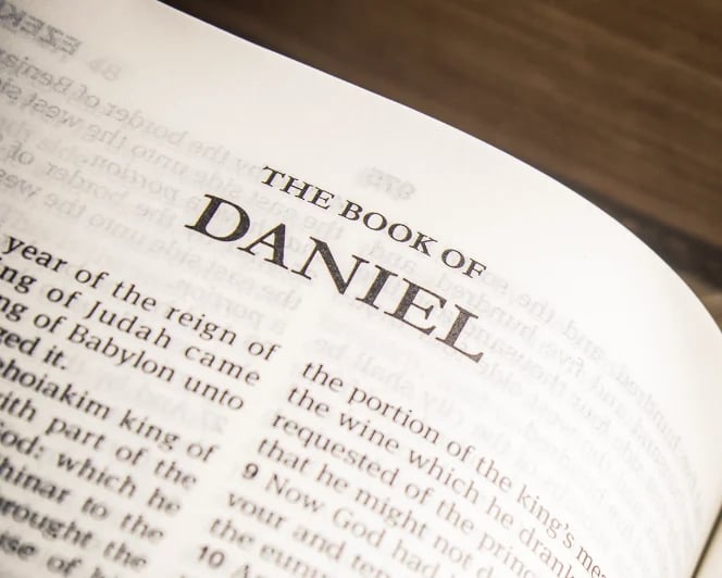 Open Bible page showing the title The Book of Daniel with text visible on white paper against a dark wooden surface