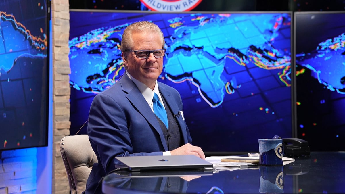 Man in dark suit and glasses seated at desk with world map display in background